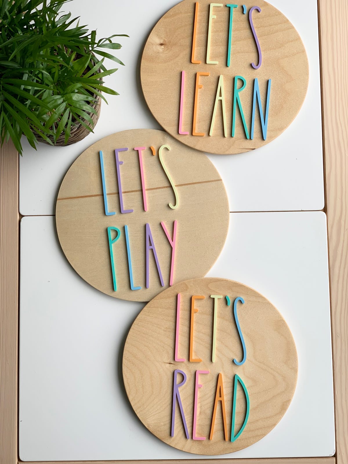 Set of three 10-inch round wooden signs with colorful pastel letters spelling Let's Play, Let's Read, and Let's Learn
