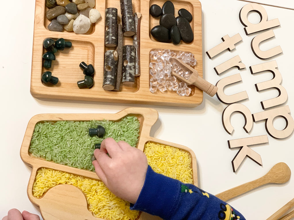Natural wooden dump truck sensory tray filled with colored rice and a child’s hand playing next to a divided tray with stones and sticks