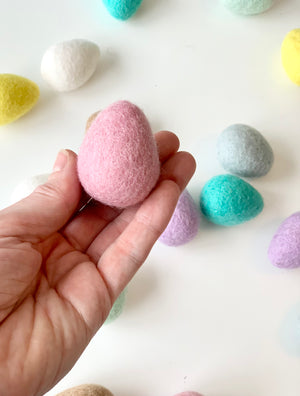 Hand holding single pink felt Easter egg with scattered pastel-colored wool eggs on white background