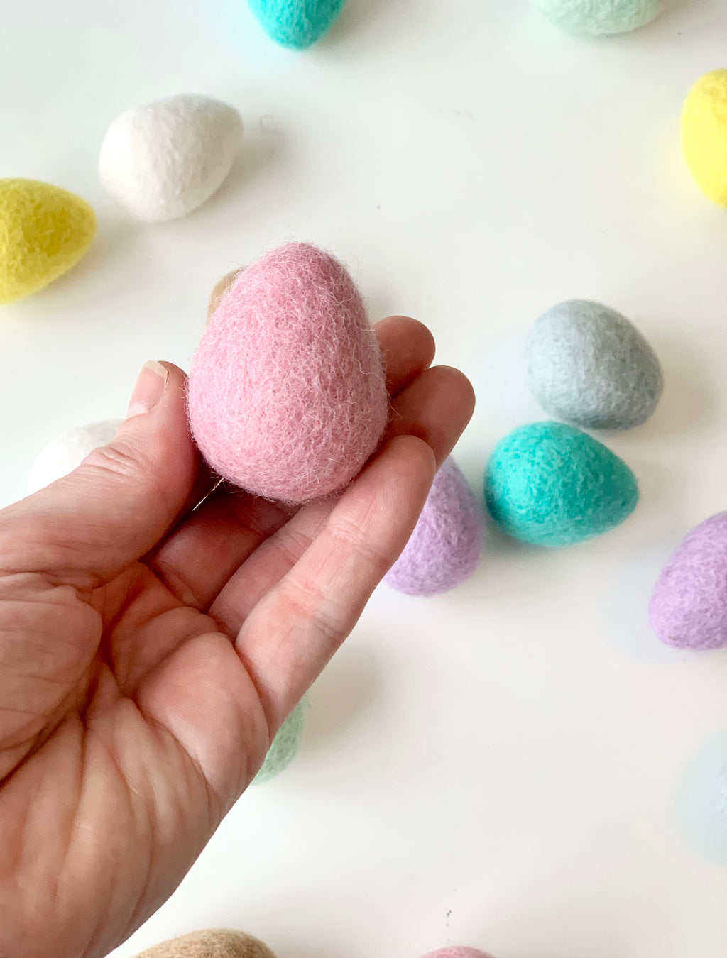 Hand holding single pink felt Easter egg with scattered pastel-colored wool eggs on white background