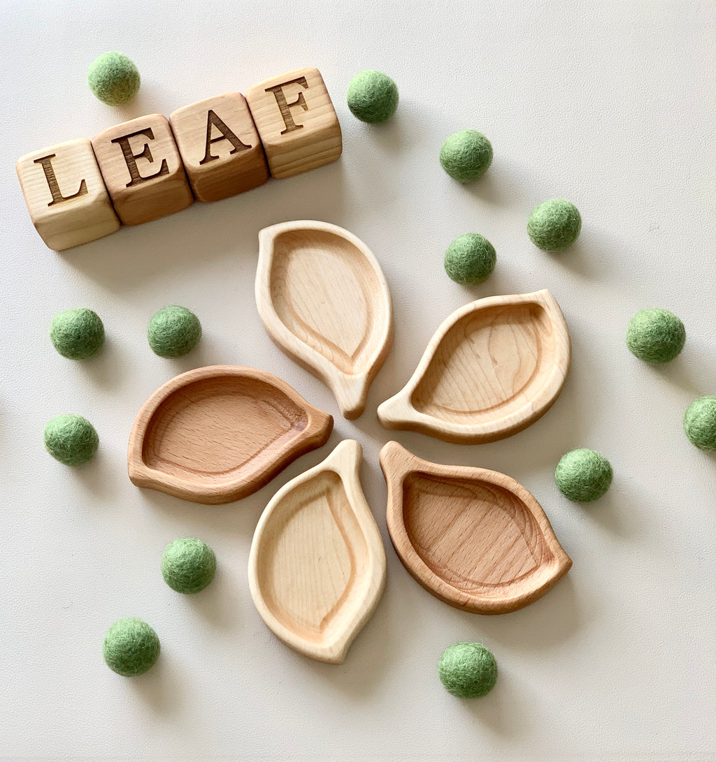 Set of small wooden leaf-shaped sensory trays with natural green felt balls and wooden blocks spelling leaf on white surface