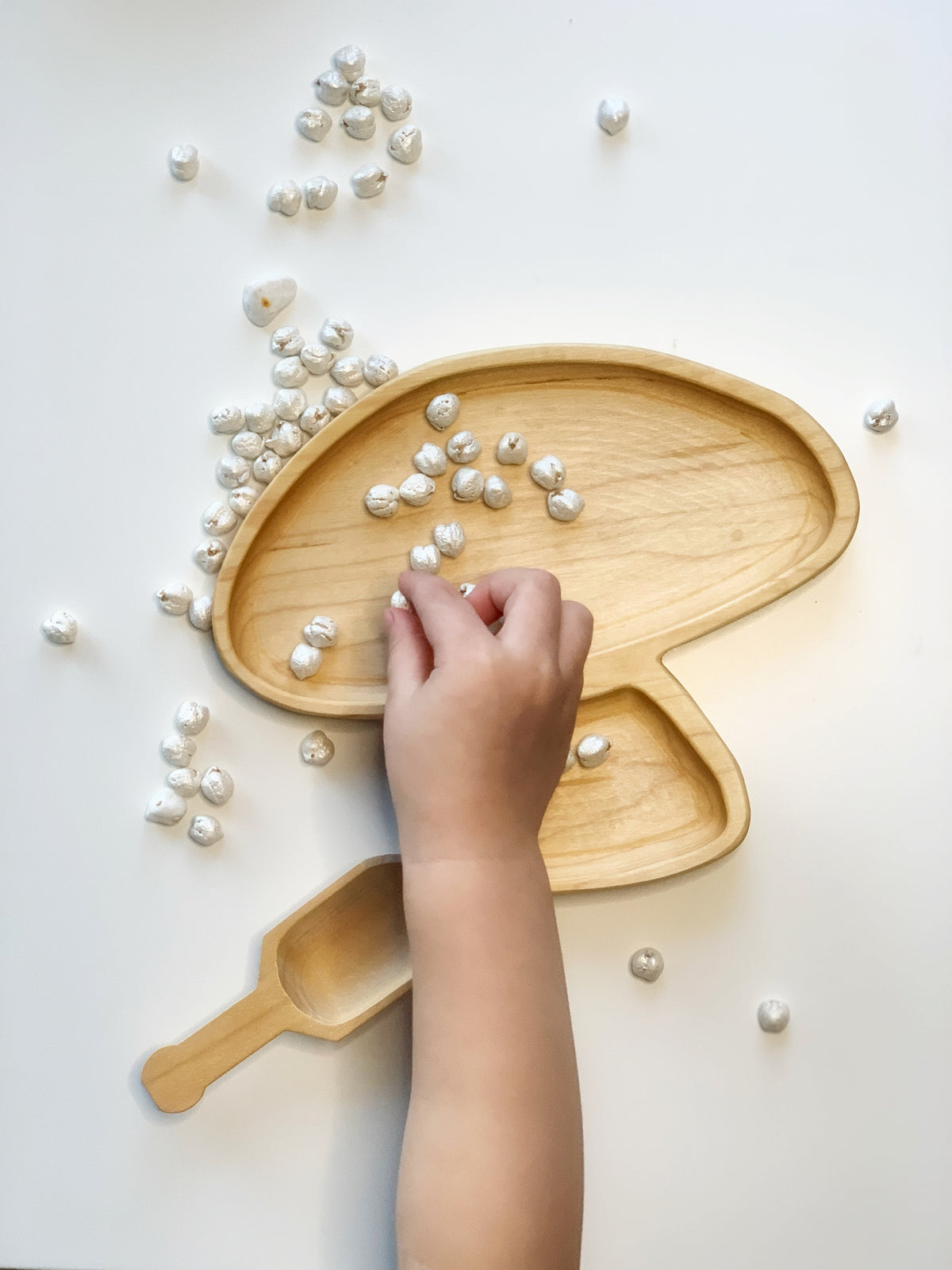 Natural wooden mushroom-shaped plate and small scoop with white pebbles scattered on white surface and child’s hand arranging pebbles