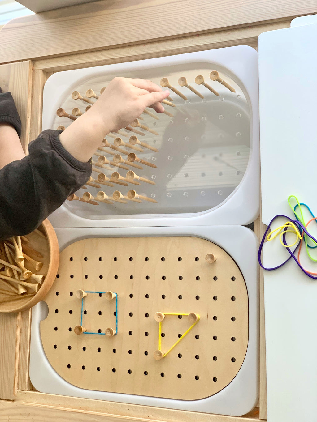 Child’s hand placing wooden peg into small hole geometric peg board with rubber band shapes on natural wood surface