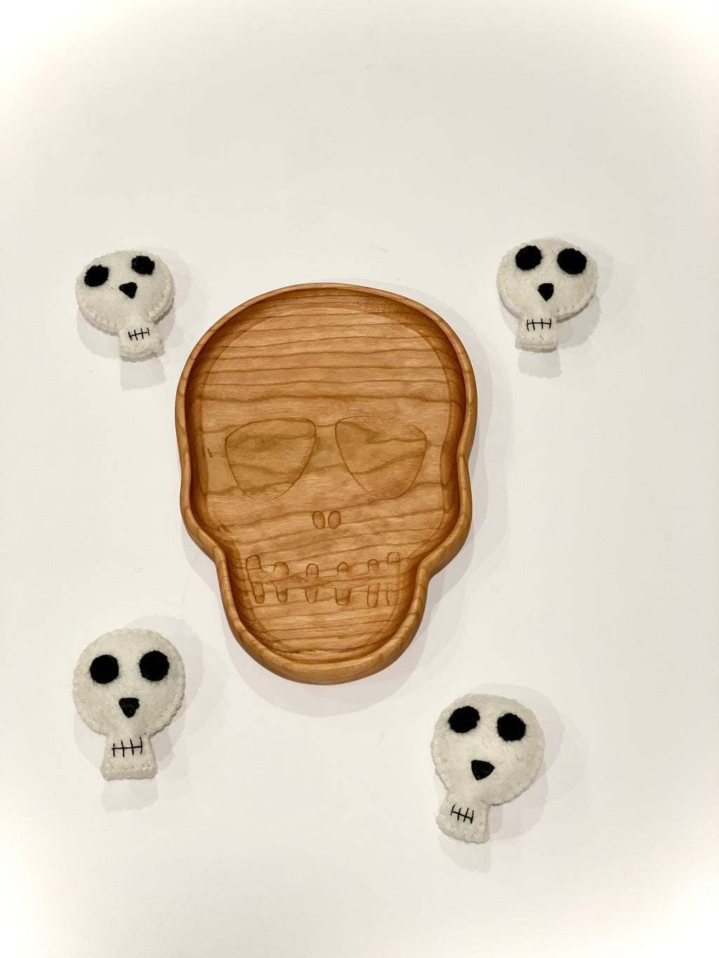 Natural wooden skull-shaped plate with engraved features surrounded by four small white felt skull toys on white background