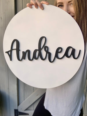 Round white 3D name sign with black cursive letters spelling Andrea held by person in light gray shirt against wooden background