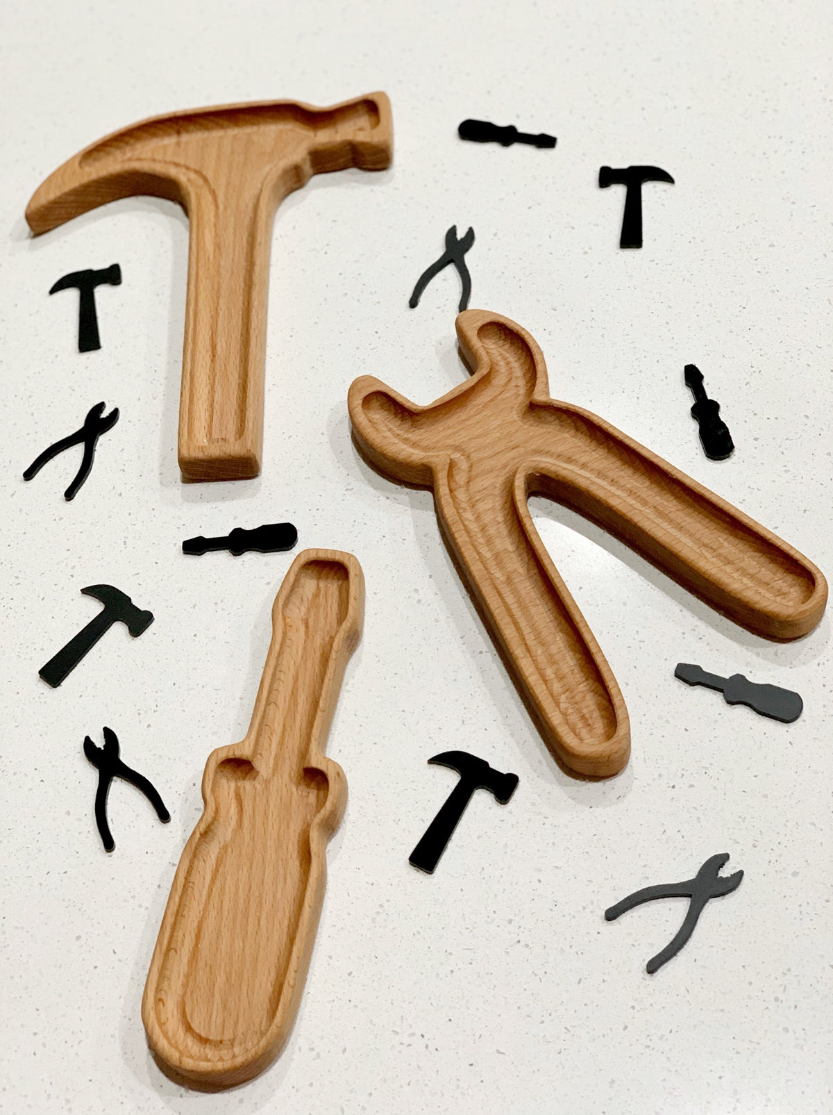 Wooden tool-shaped sensory trays in hammer, pliers, and screwdriver shapes with small black tool silhouettes on white surface