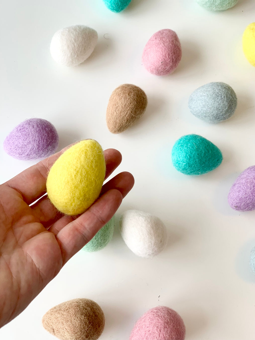 Hand holding one yellow felt Easter egg with scattered pastel colored felt eggs on white surface