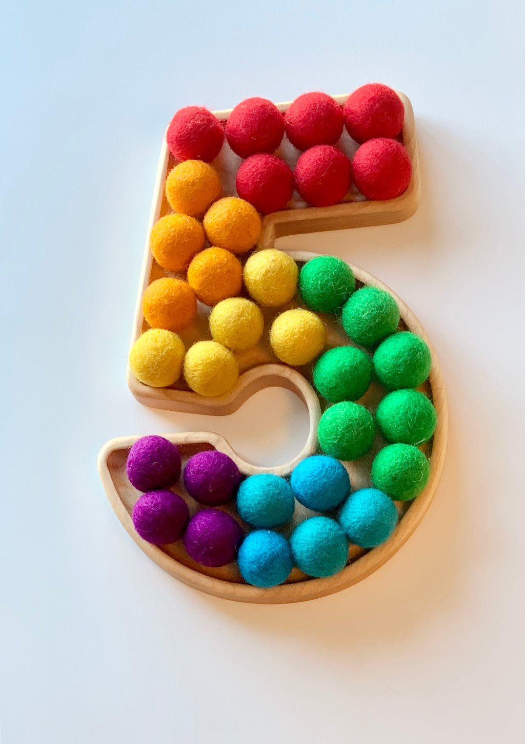 Wooden sensory tray shaped as number 5 filled with colorful felt balls in rainbow colors on white background