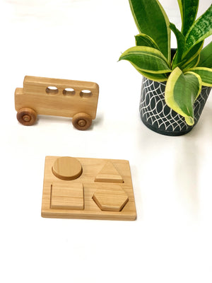Natural wooden shape puzzle with circle, triangle, square, and hexagon pieces next to wooden toy bus and potted plant on white background