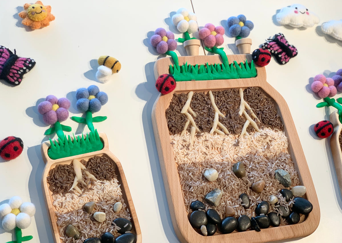 Large wooden sensory tray shaped like a jar filled with layered seeds and beans with felt flowers and ladybug decorations