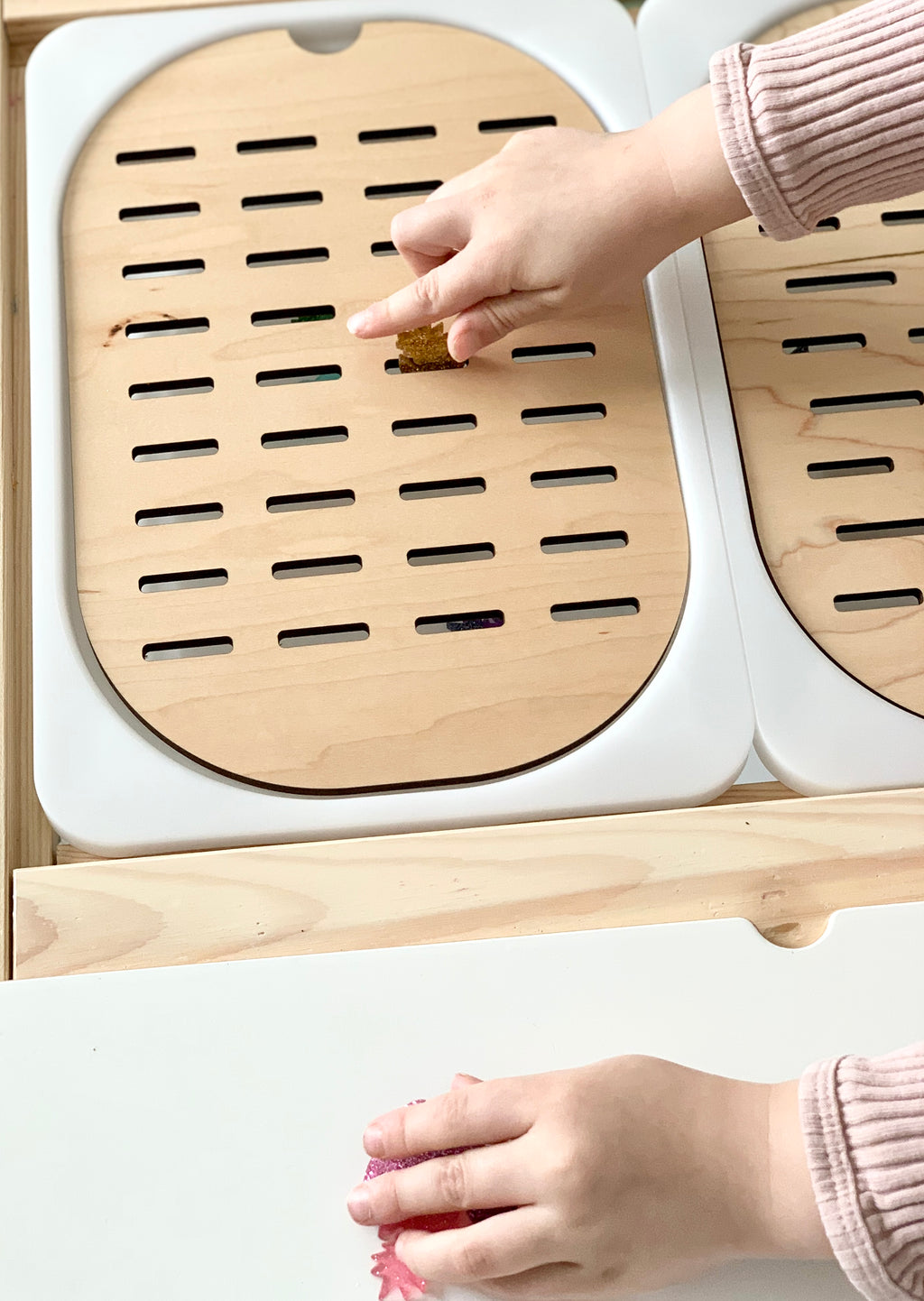 Child's hands placing objects through wooden slot sorter Flisat table top insert with white frame on wood surface