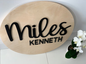 Large 3D wooden round name sign with black cursive and block letters on natural wood background near white orchid plant