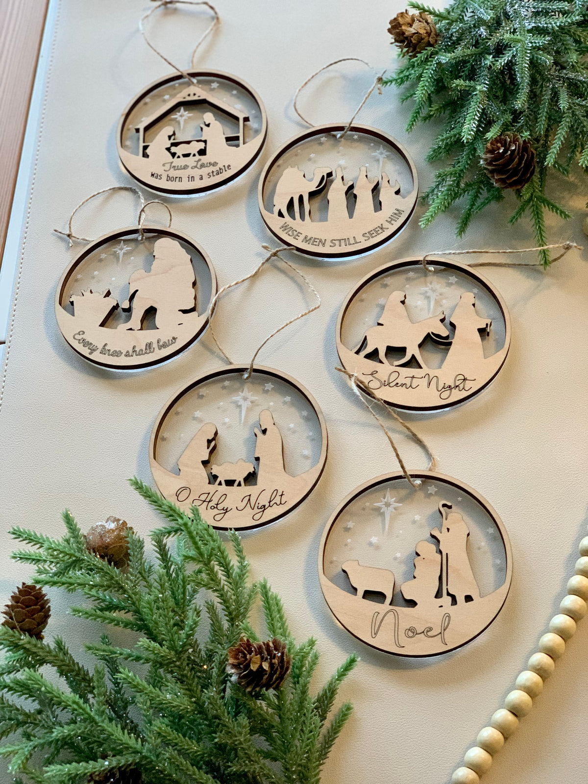 Two-layer wood and acrylic nativity Christmas ornaments with biblical scenes and festive inscriptions on white surface with greenery