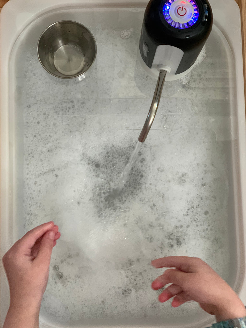 Top view of play kitchen sink insert with foamy water, running faucet, metal cup, and child’s hands in sink