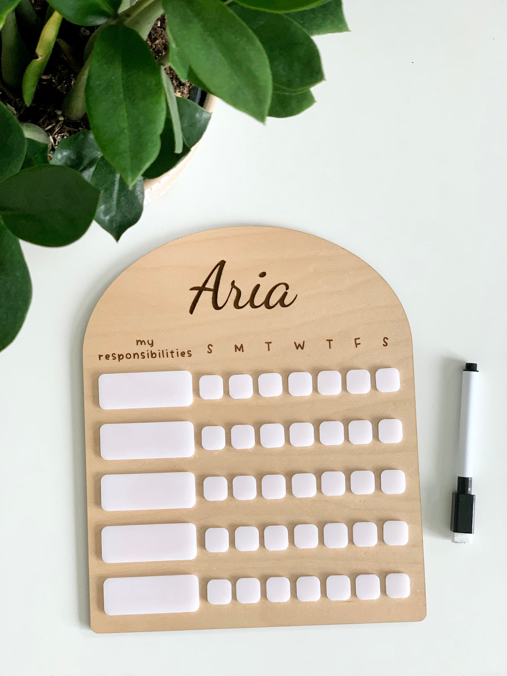 Personalized wooden chore chart with white acrylic dry erase pieces and black marker on white surface next to green plant