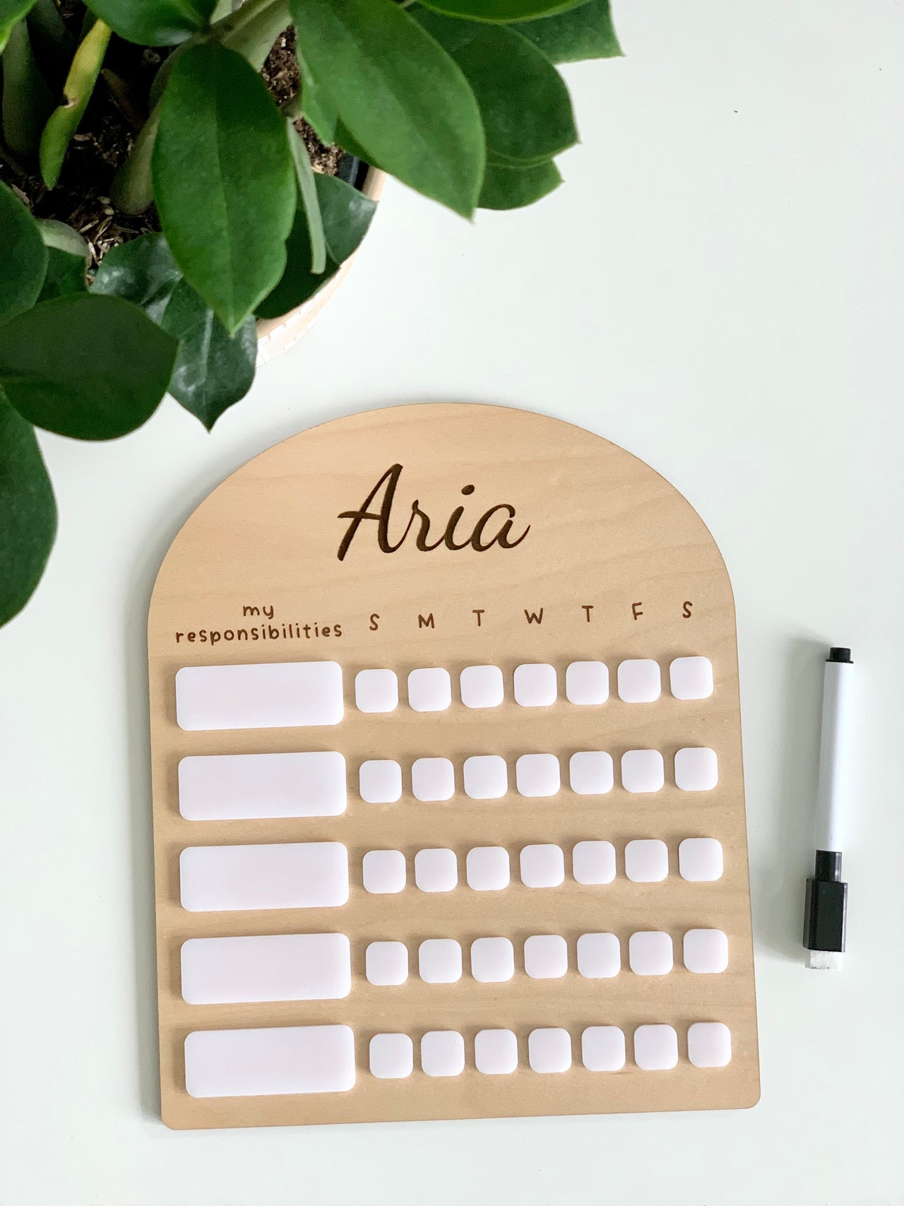 Personalized wooden chore chart with white acrylic dry erase pieces and black marker on white surface next to green plant