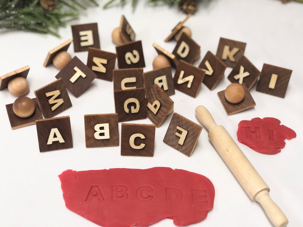 Natural wooden alphabet play dough stampers with engraved capital letters scattered beside red play dough and wooden rolling pin