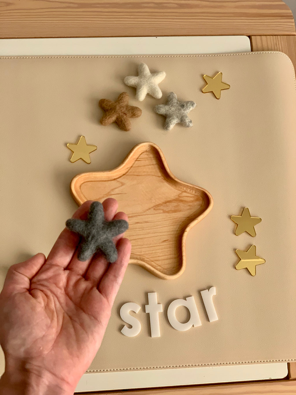Hand holding a gray felt star above a wooden star-shaped tray with other felt stars and white letter blocks spelling star
