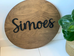 Round wooden name sign with black cursive letters on natural wood background beside green potted plant