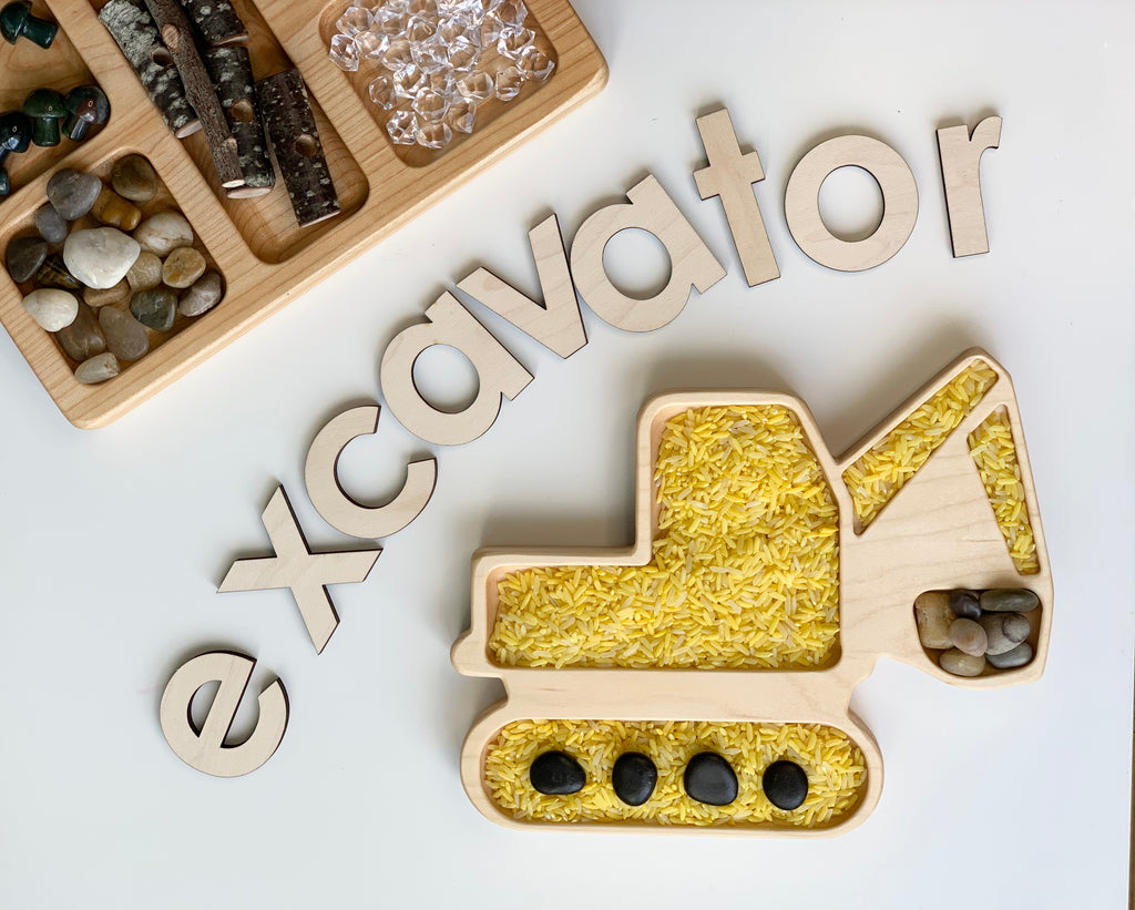 Wooden excavator-shaped sensory tray filled with yellow rice and small stones beside wooden letters spelling excavator on white surface