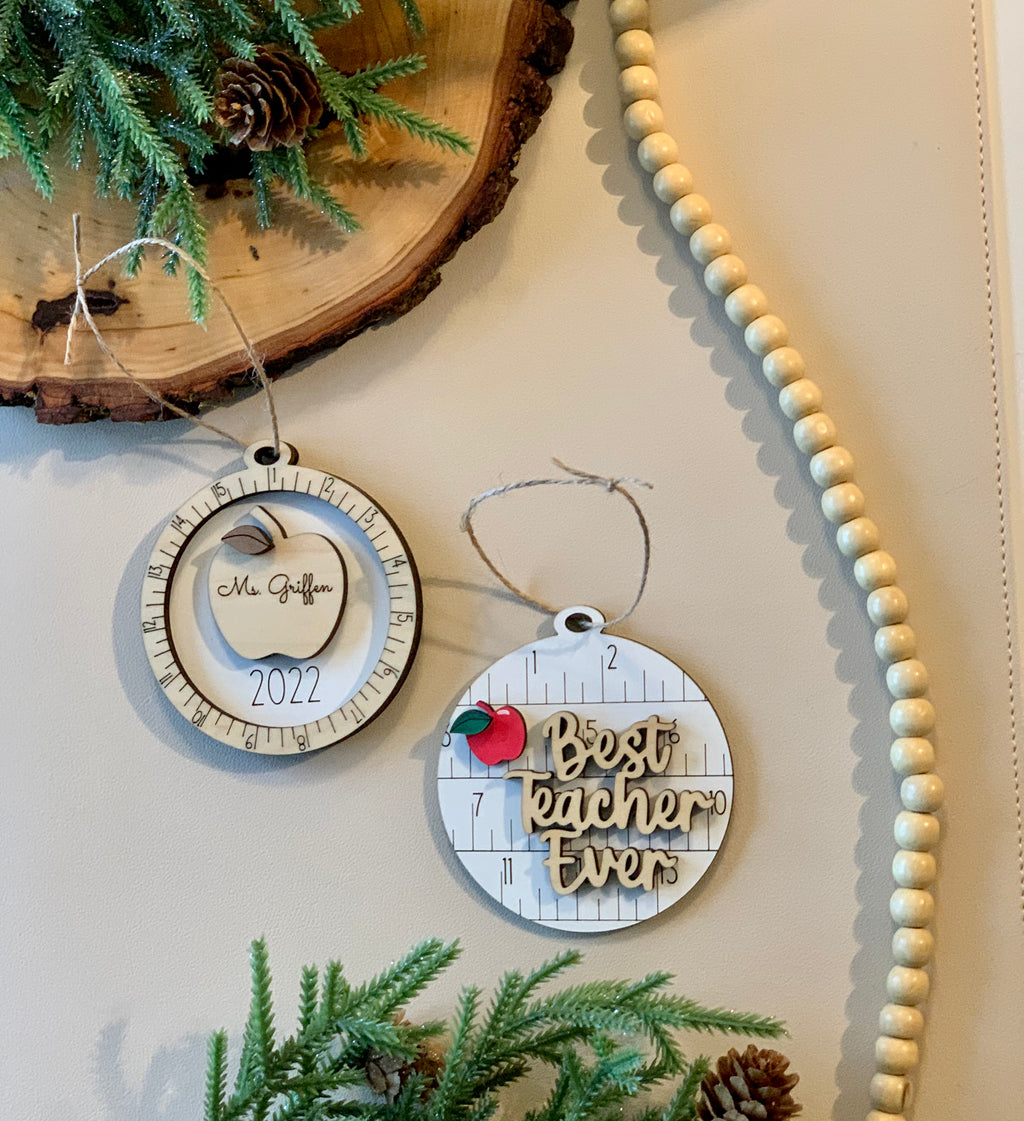 Two round wooden Christmas ornaments with measuring tape design, one engraved with apple and name, the other with "Best teacher ever" text on white surface
