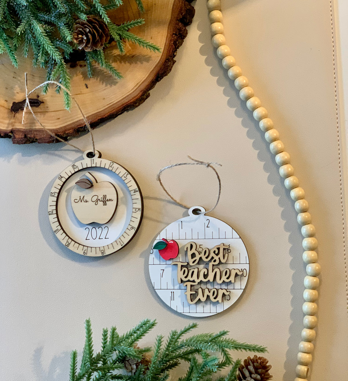 Two round wooden Christmas ornaments with measuring tape design, one engraved with apple and name, the other with "Best teacher ever" text on white surface