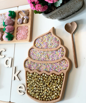Wooden cupcake-shaped sensory tray with colorful rice and gold beads beside heart-shaped wooden spoon and pink flowers