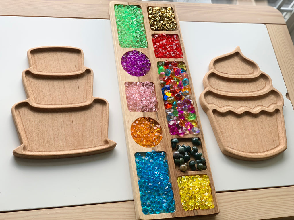 Wooden cake and cupcake shaped sensory trays alongside a divided wooden tray filled with colorful gem beads