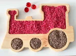Natural wooden train-shaped sensory tray filled with pink and brown colored rice and three red gem-like beads on white surface