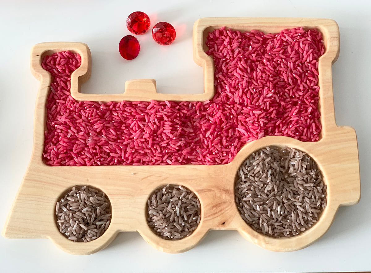 Natural wooden train-shaped sensory tray filled with pink and brown colored rice and three red gem-like beads on white surface