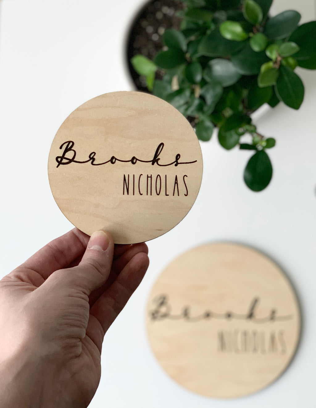 Hand holding small round maple wood baby name announcement disc with cursive and block engraved letters and plant background