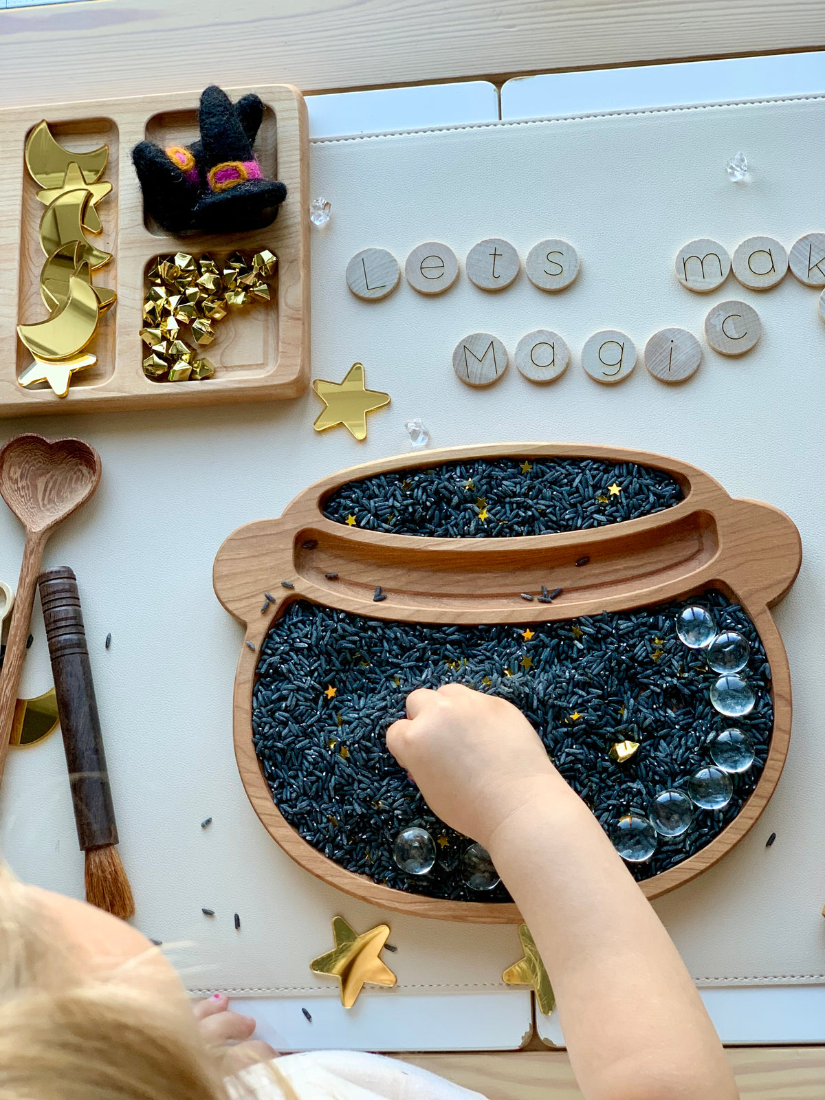 Wooden cauldron-shaped sensory bin filled with dyed black rice and gold star confetti with child's hand playing