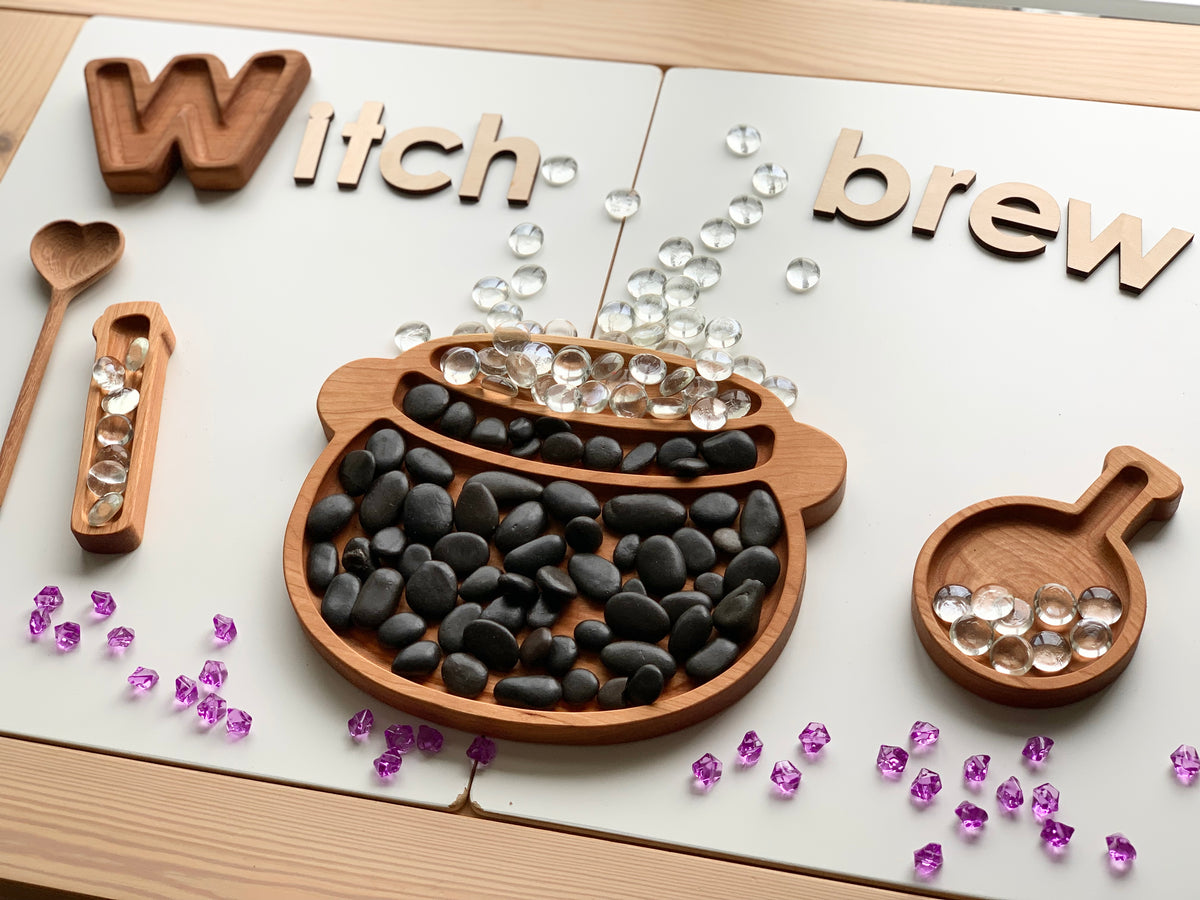 Wooden sensory tray shaped like a cauldron filled with black stones and clear gems under wooden letters spelling Witch brew