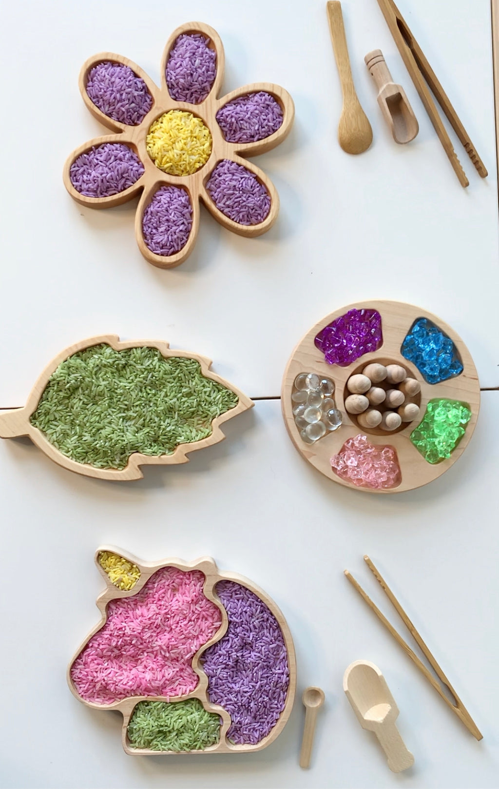 Wooden sensory trays shaped like flower, leaf, and unicorn filled with colored rice and beads on white surface