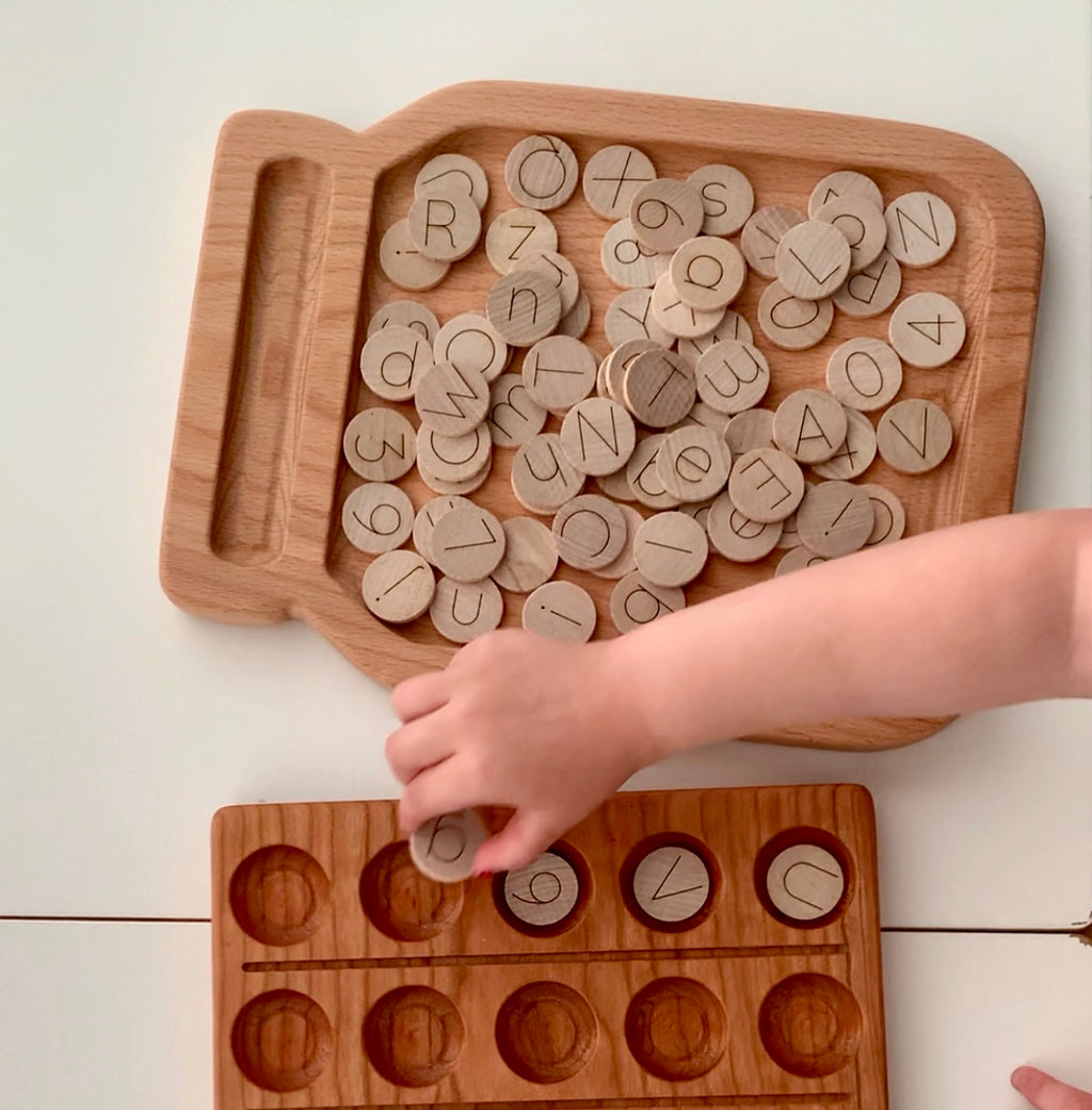 Wooden 1-inch alphabet and number discs with simple laser engraved letters and numbers on natural wood tray with child’s hand