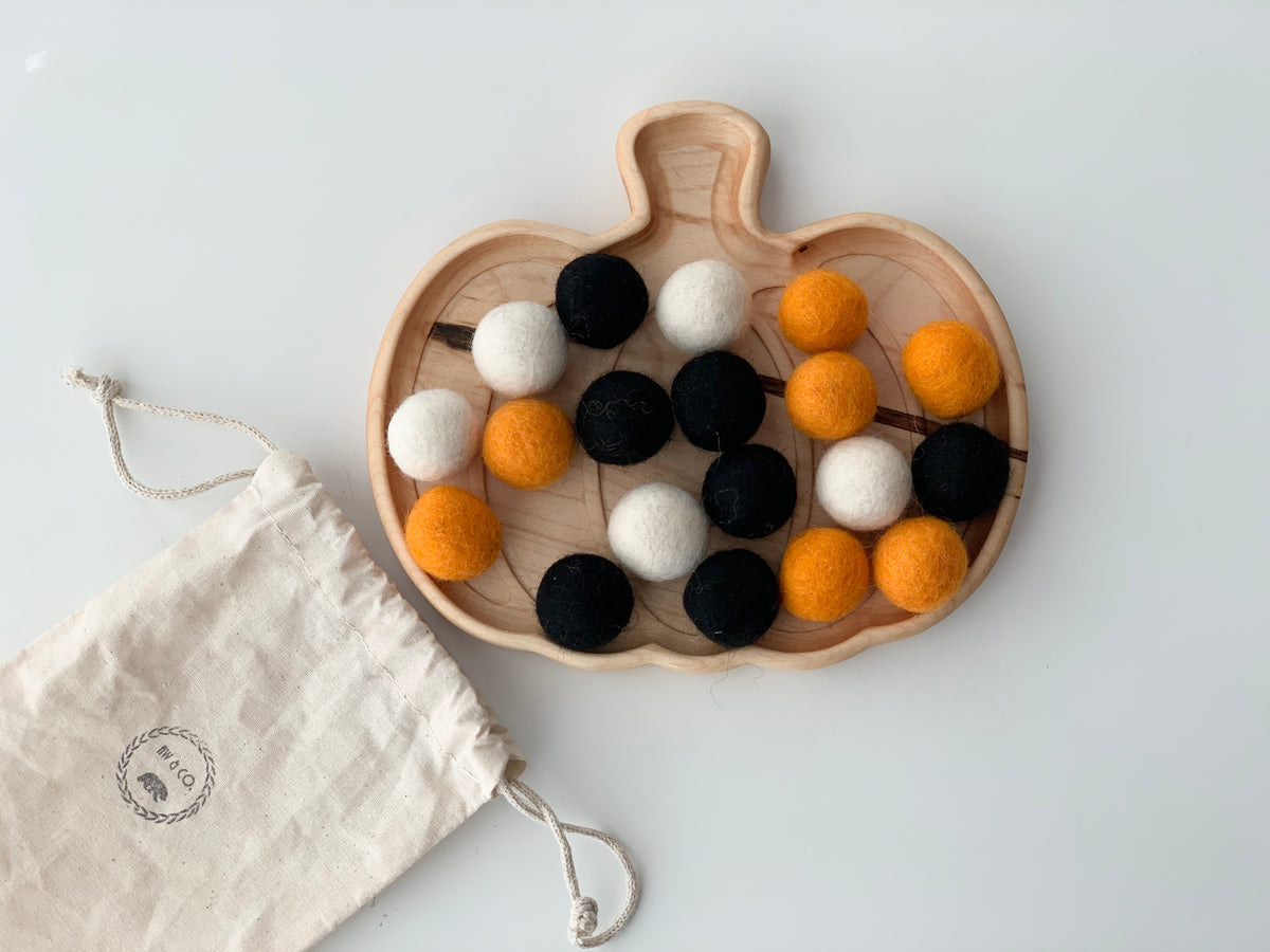 Assorted black, orange, and white felt balls displayed on wooden pumpkin tray next to white drawstring bag on white surface