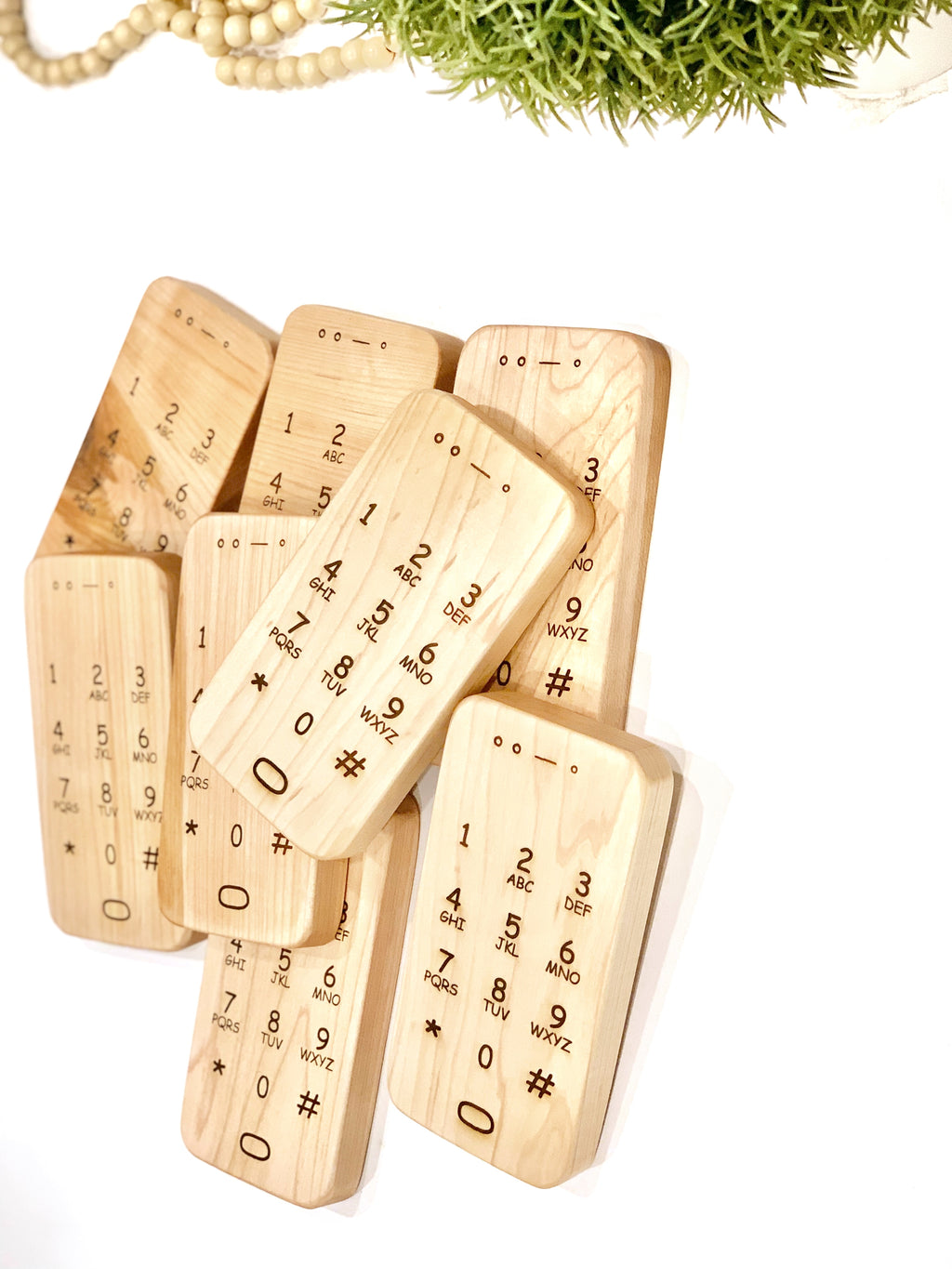 Natural wooden toy phones with engraved number pads arranged overlapping on white surface with green plant above