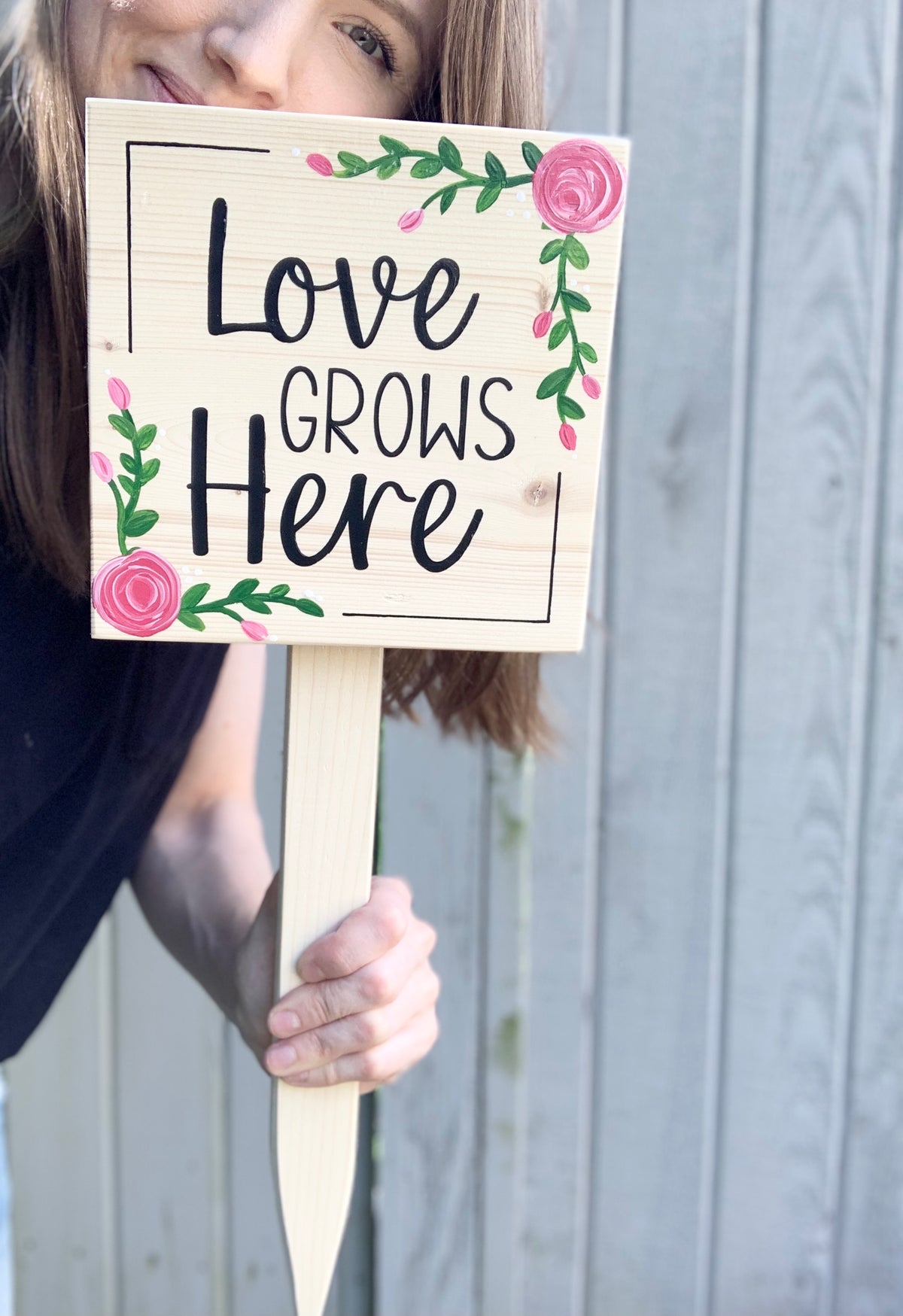 Small square wooden garden sign with "Love Grows Here" text and pink floral painted decorations on natural wood background
