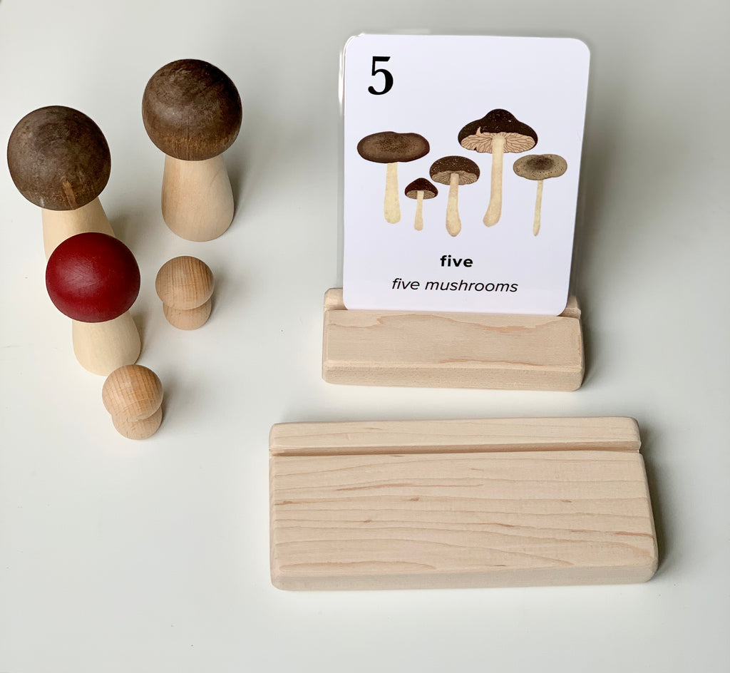 Natural unfinished wooden flash card holder with a card showing five mushrooms and wooden mushroom toys nearby on white surface