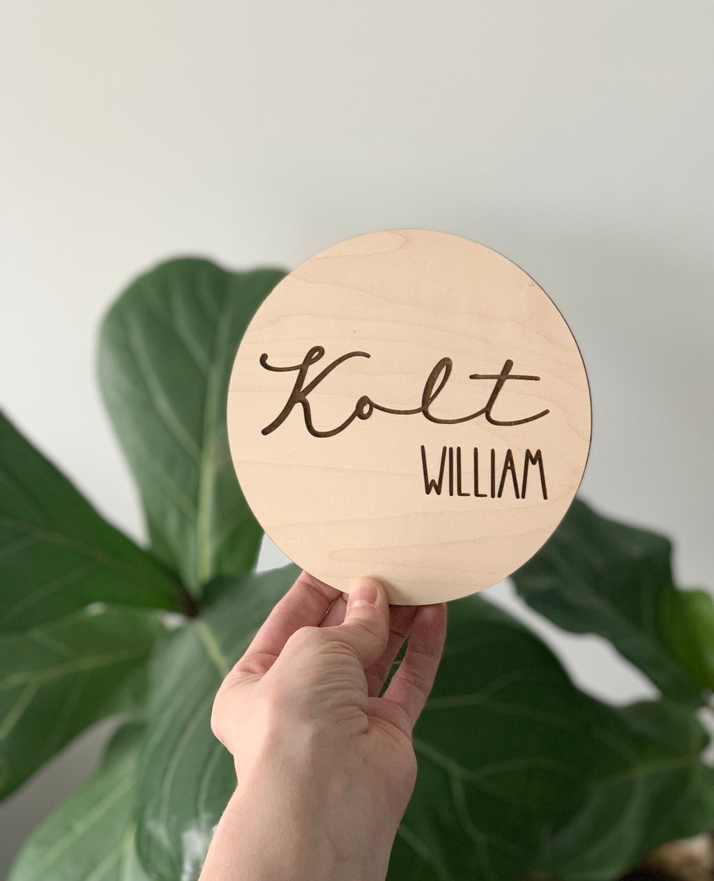 Hand holding personalized 4-inch maple wood baby name announcement disc with Kolt William engraved text