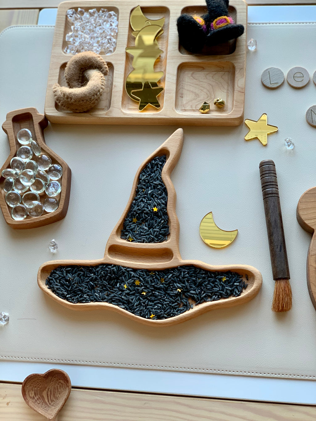 Natural wooden witch hat sensory tray filled with black rice and gold star confetti on white surface with accessories