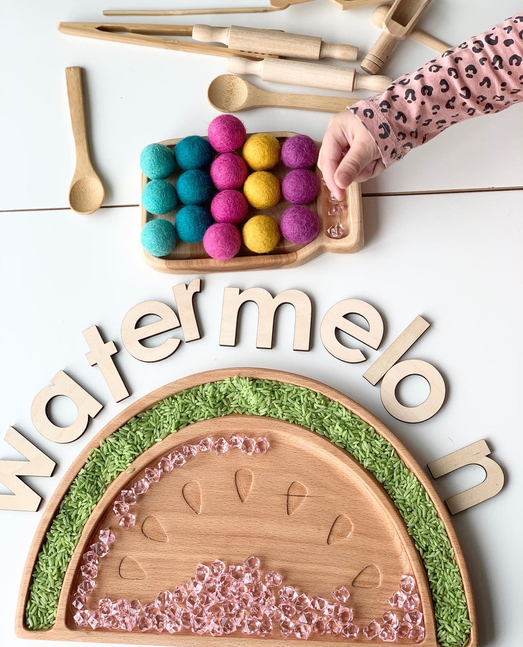 Wooden watermelon-shaped sensory tray with pink beads, green rice, colorful felt balls, and wooden letters spelling watermelon