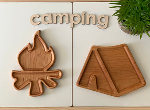 Two natural wooden sensory trays shaped like a campfire and a tent with wooden letters spelling camping on white surface