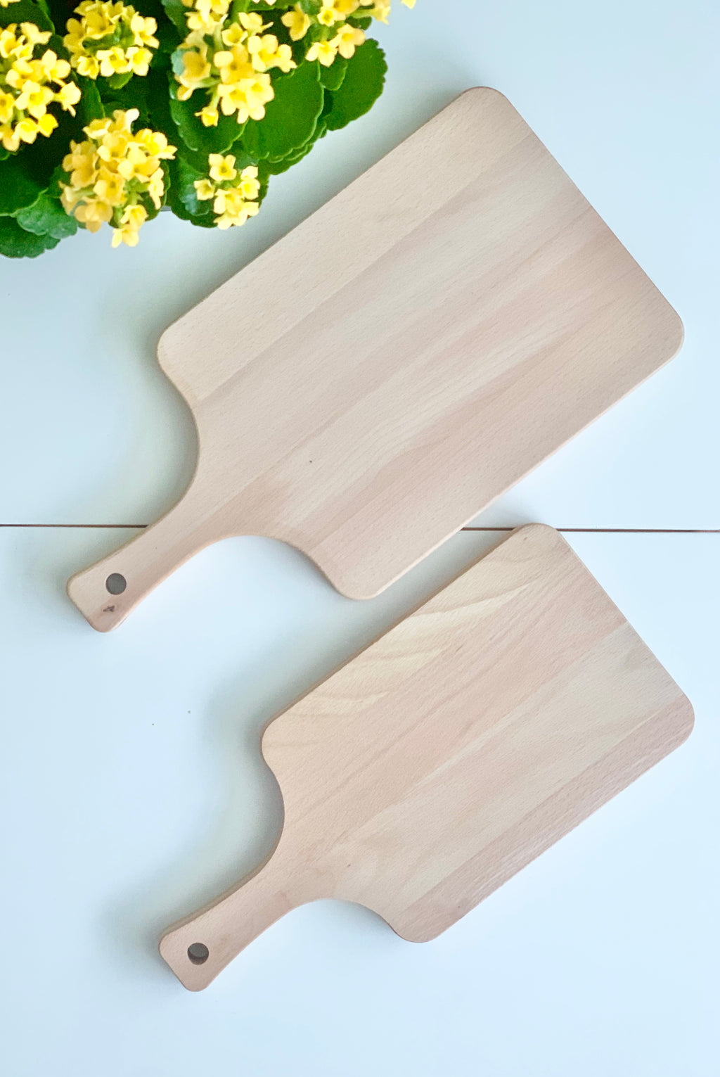 Two natural wooden personalized cutting boards with handles placed on white surface beside yellow flowering plant