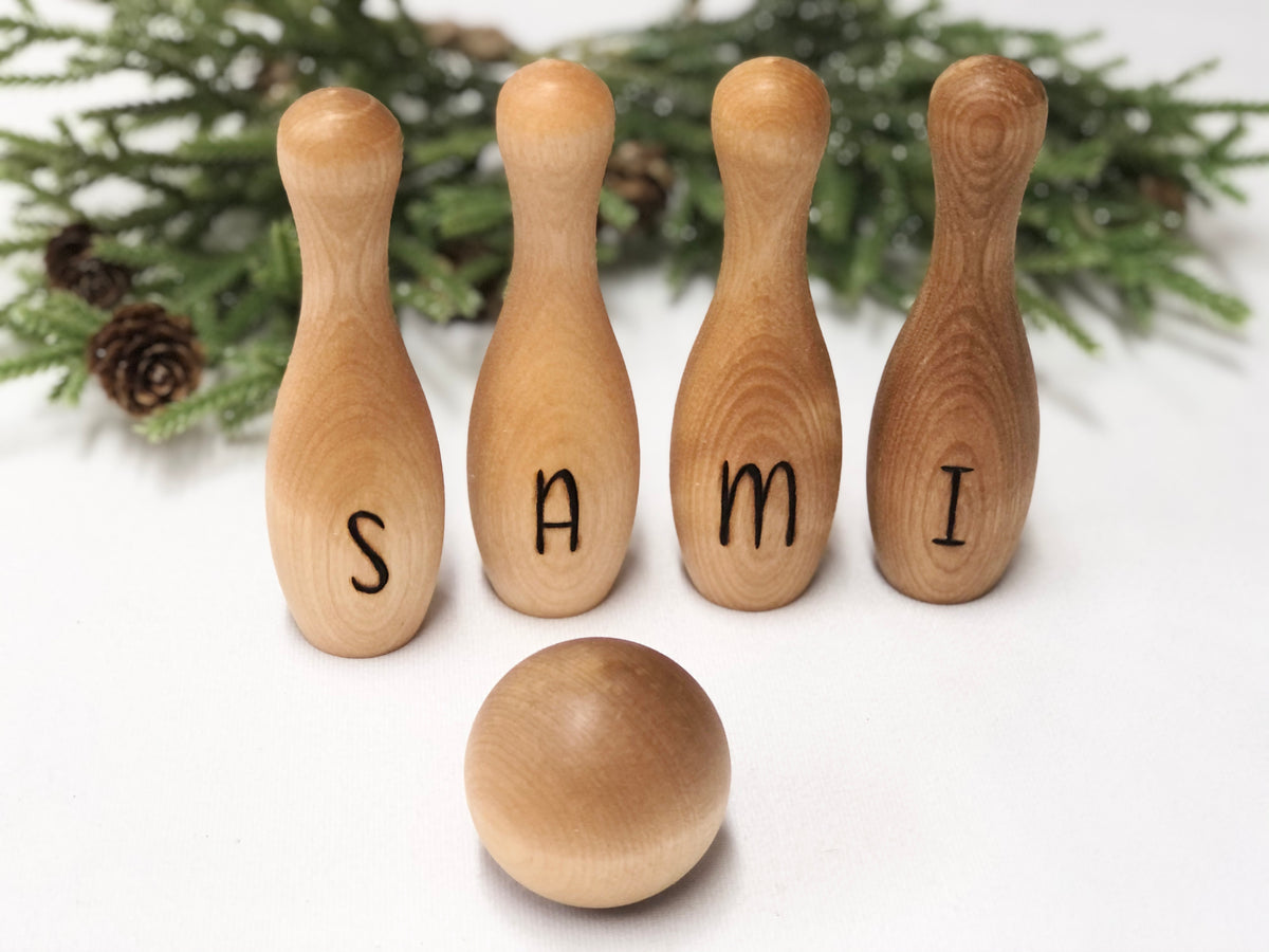 Personalized wooden bowling pins spelling SAMI with a wooden ball and greenery background, toddler toy set
