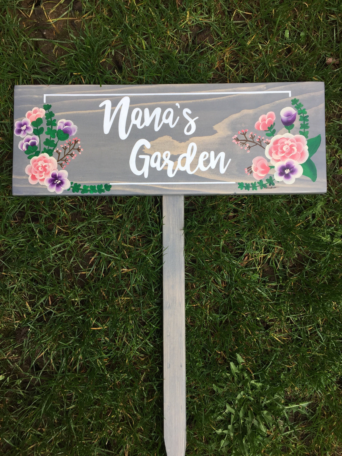 Gray wood garden sign with white cursive letters saying Nana’s Garden and pink and purple floral accents on grass background