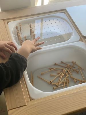 Child’s hands arranging small wooden pegs into a white geometric light board insert on a natural wood table top