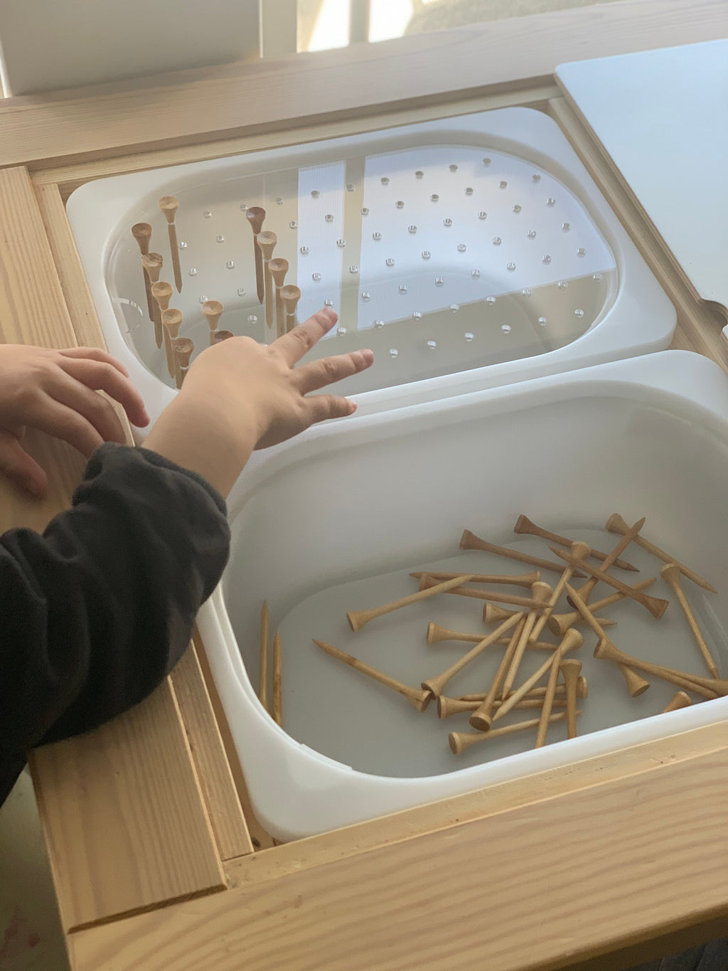 Child’s hands arranging small wooden pegs into a white geometric light board insert on a natural wood table top