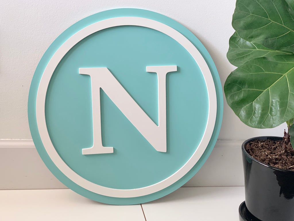 Round 3D monogram wooden sign with white letter N on teal background and white border leaning on white wall next to plant
