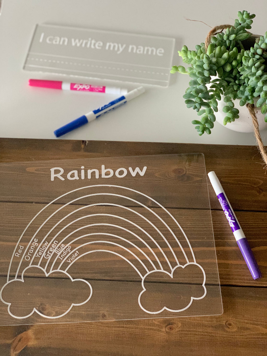 Clear acrylic tracing board engraved with rainbow and color names, dry erase markers, and plant on wooden table
